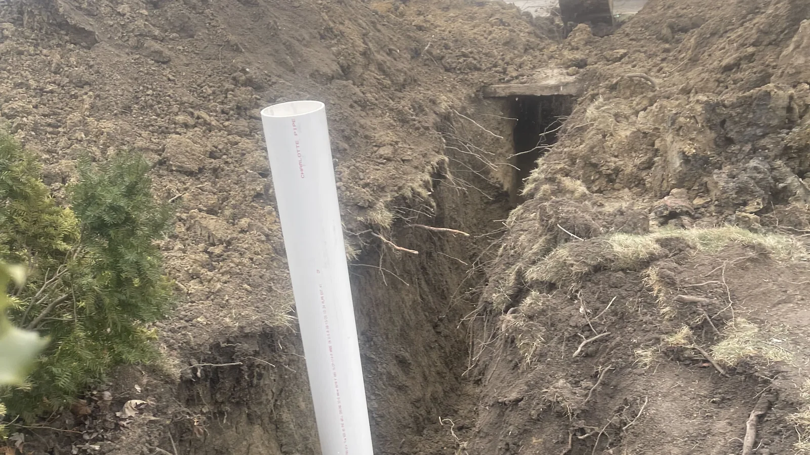 Deep trench with a new white PVC cleanout riser and spoil piled along both sides, underground utility work