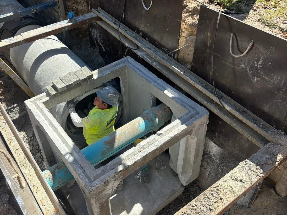 Crew member inside a precast structure fitting a pipe connection in a shored excavation, underground utility work