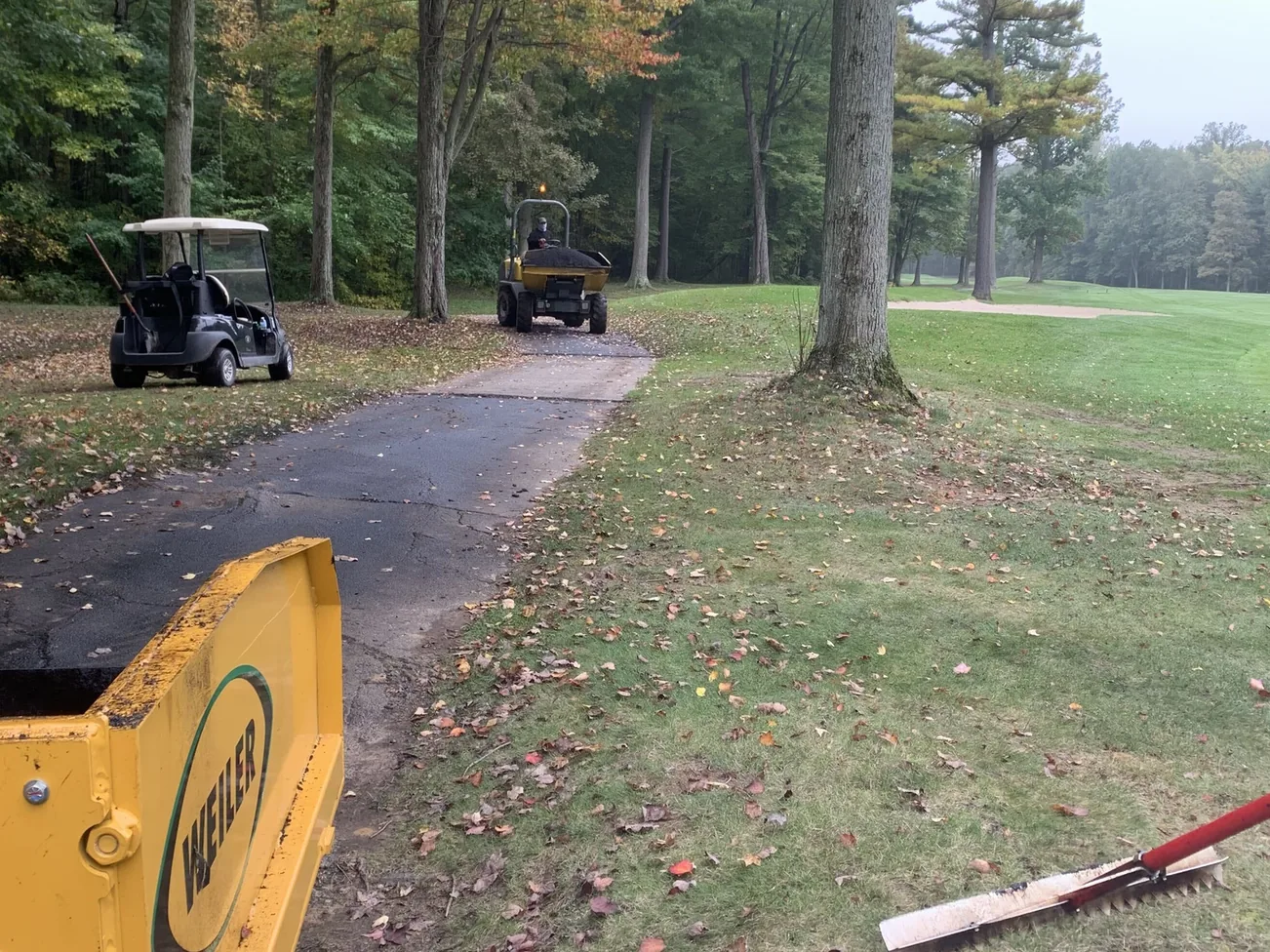 Fresh asphalt cart path paved through a wooded golf course with the paver screed in the foreground, asphalt paving