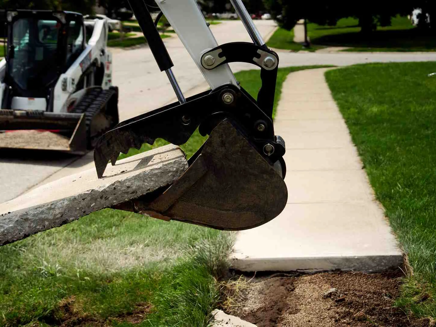 Mini excavator thumb lifting a broken slab of an old concrete walk beside a residential street, concrete removal