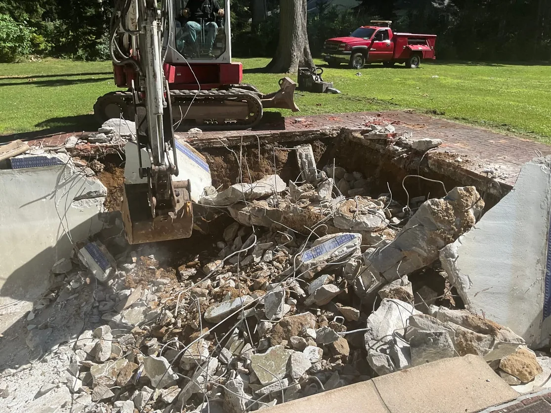 Mini excavator breaking out an old inground pool, the broken shell, tile and rebar piled in the hole, pool demolition