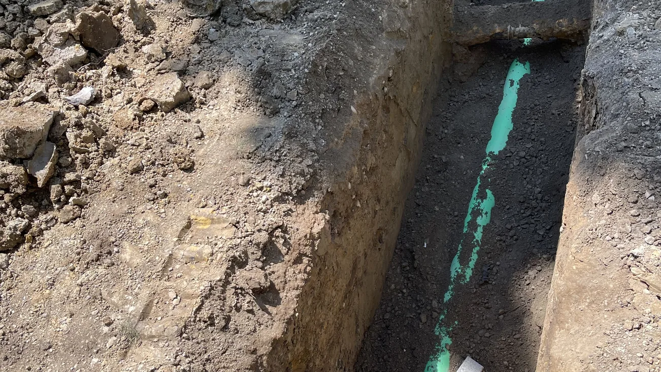 Trench with a new green PVC sewer lateral beside old pipe stubs, sewer line work