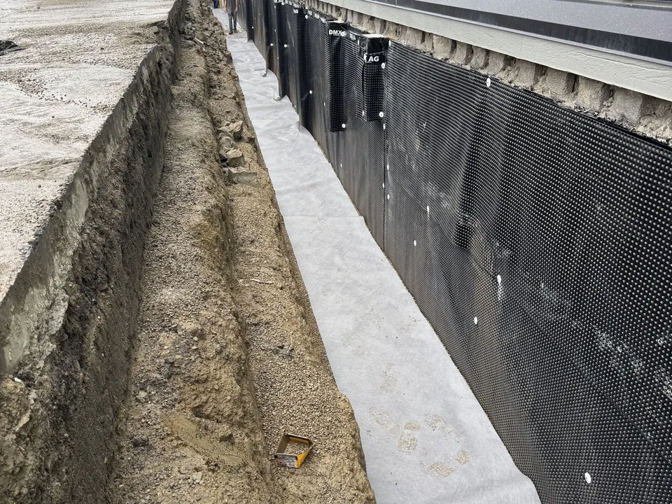 Open trench along a building foundation with black waterproofing membrane and drainage board, foundation waterproofing