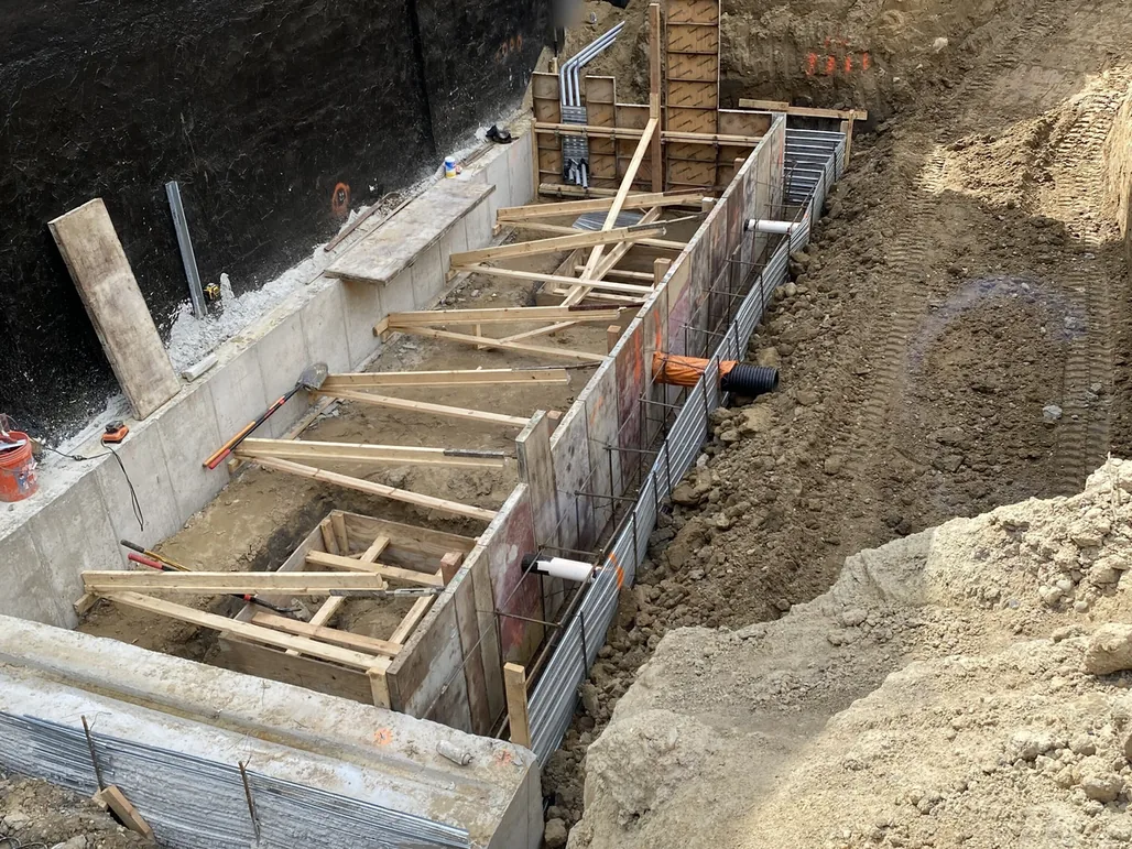 Black waterproofing membrane on a foundation wall beside new forms and rebar for an adjoining wall, foundation waterproofing