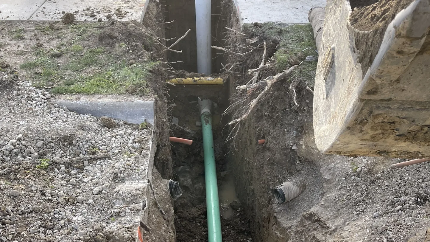 Excavator bucket at an open trench with new green PVC pipe laid in it beside a sidewalk, septic system installation and underground work