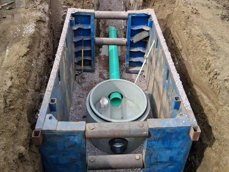 Trench box set in a long open trench with a precast manhole base and new green PVC pipe, construction project management