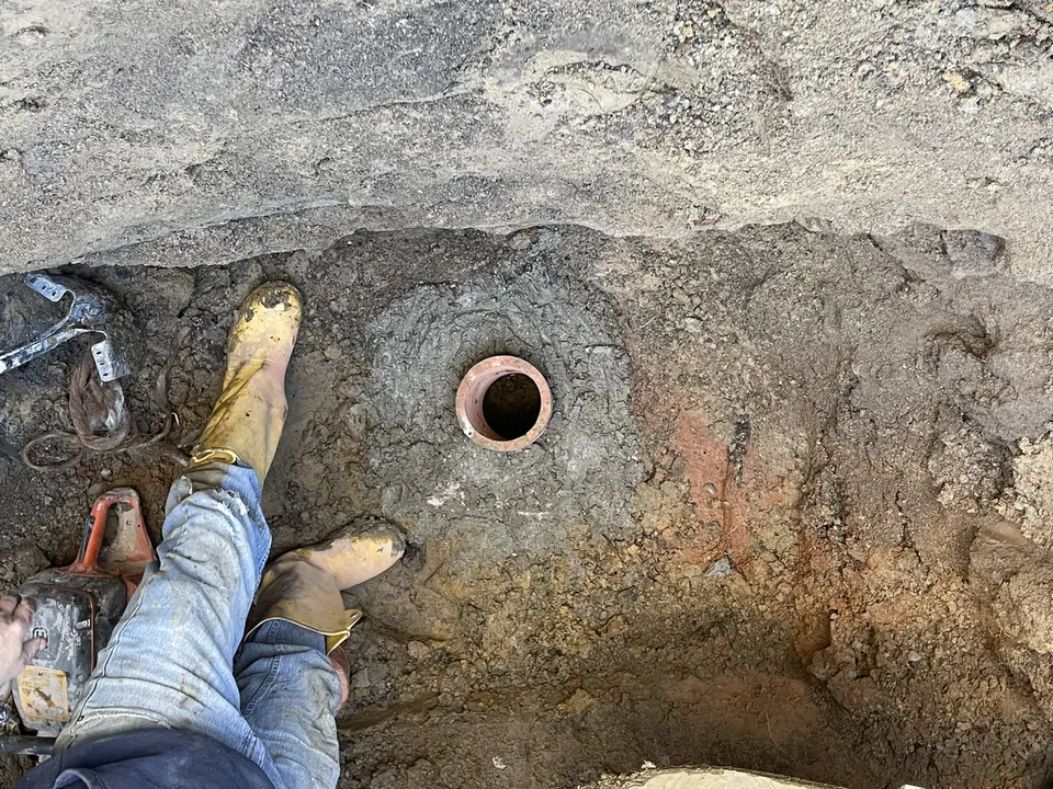 Old clay pipe opening exposed in the floor of a dug pit beside the boots of a worker and a power saw, septic system repair