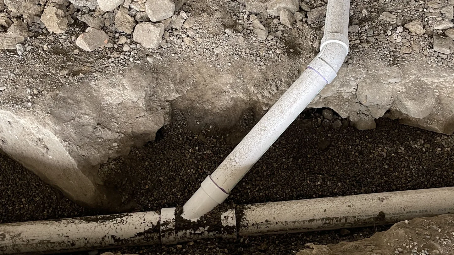 New white PVC line angled down through stone fill and tied into a drain line in a gravel trench, underground utility work
