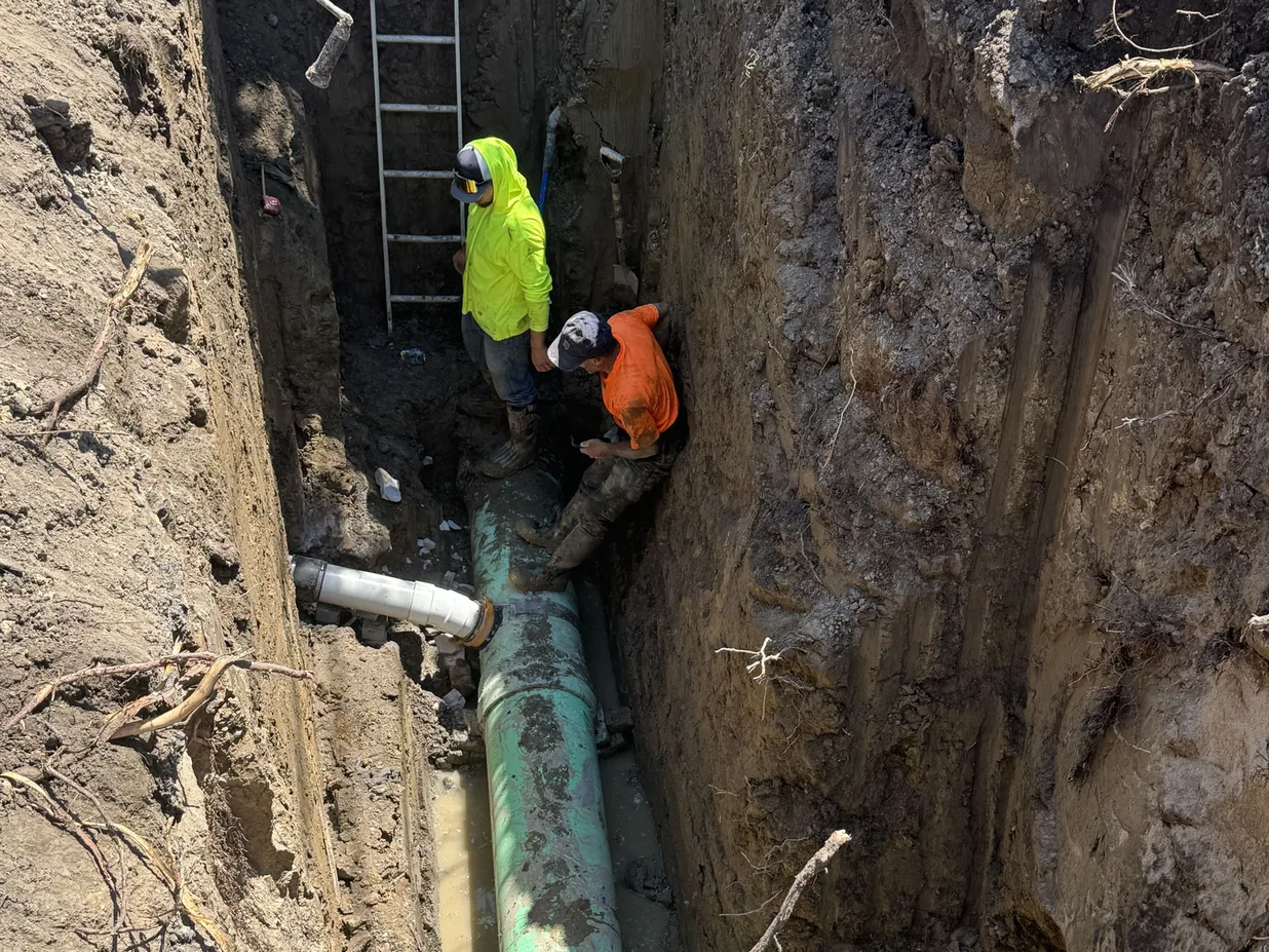 Two crew members setting a green PVC sewer main at the bottom of a deep trench, sewer line replacement
