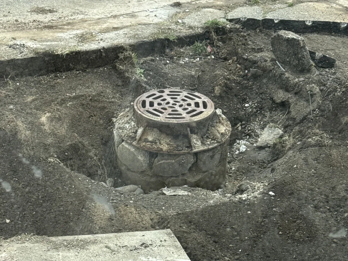 Round storm drain grate set in a fresh concrete collar beside old pavement, storm drainage
